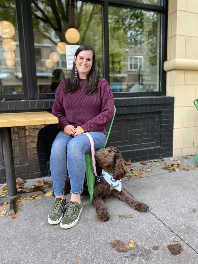 A woman in jeans and a maroon top sits outside at a table with a brown doodle under her chair, looking up at her.