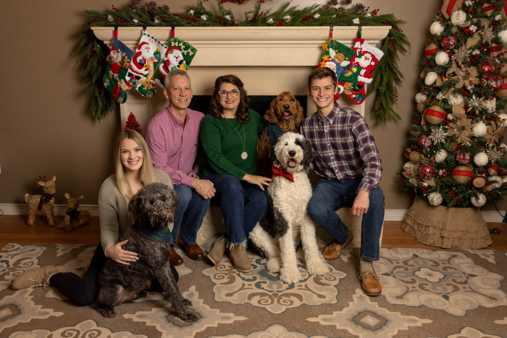 A family in front of a fireplace with a Christmas tree and decorations surrounding them. There are four people and three dogs.